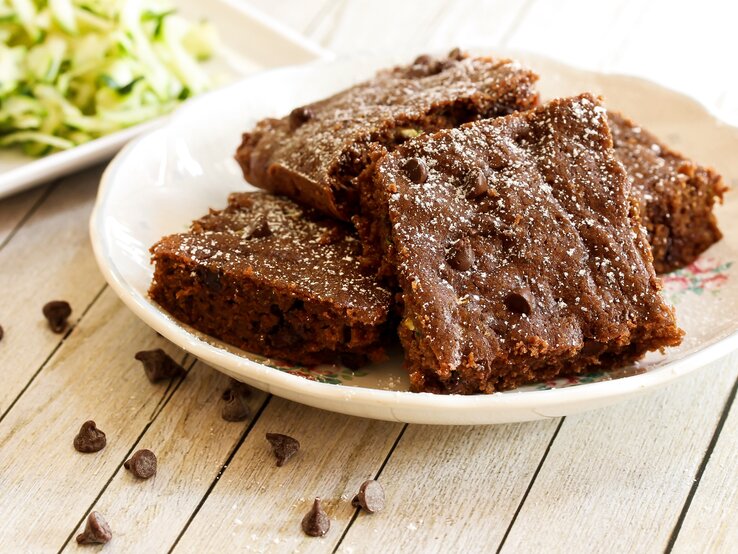 Ein weißer Teller mit den Zucchini-Brownies auf einem Holztisch von der Seite fotografiert. Im Hintergrund frische Zucchini unscharf zu erkennen.