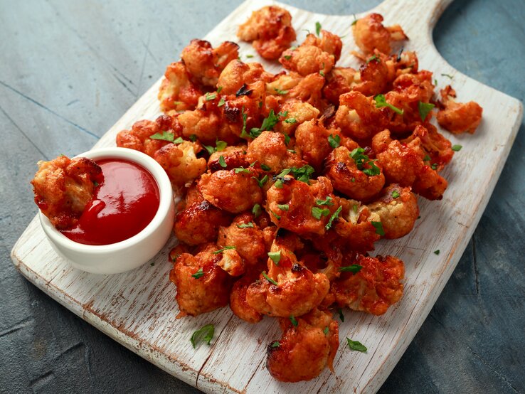 Blumenkohl Wings in scharfer Soße liegen auf einem Holzbrett auf einem grauen Untergrund. Neben den Wings steht eine kleine Schüssel mit scharfer Sauce.