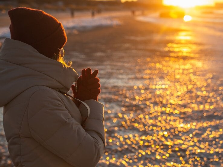 Eine Frau mit beigem Wintermantel und roter Mütze steht bei Sonnenuntergang am Strand und wärmt ihre Hände.