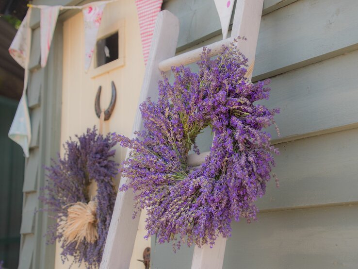 Ein großer Lavendelkranz an einer Leiter neben einer kleinen Holztür mit Hufeisen und bunten Wimpeln.