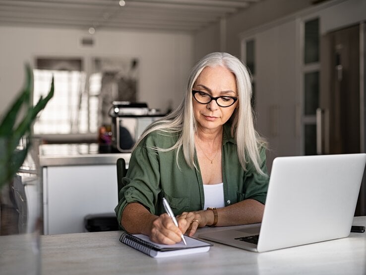 Vergesslichkeit behandeln Eine konzentrierte ältere Frau mit langen grauen Haaren und Brille sitzt an einem hellen Arbeitsplatz und schreibt etwas in ein Notizbuch. Vor ihr steht ein geöffneter Laptop. Sie trägt ein olivgrünes Hemd und wirkt, als wäre sie gerade in einer Arbeits- oder Lernumgebung zu Hause. Das Zimmer ist hell und modern eingerichtet, mit einer Küche im Hintergrund, was auf ein häusliches Büro hindeutet. Das natürliche Licht und die Pflanze auf dem Tisch schaffen eine angenehme und ruhige Arbeitsatmosphäre.