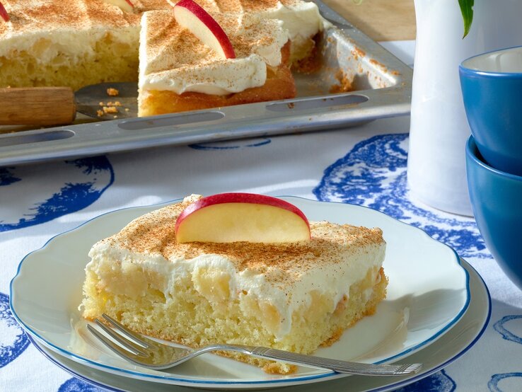 Ein Stück Apfelkuchen mit Schmandhaube auf weiß-blauem-Hintergrund. Dahinter das Kuchenblech und rechts davon Tassen.