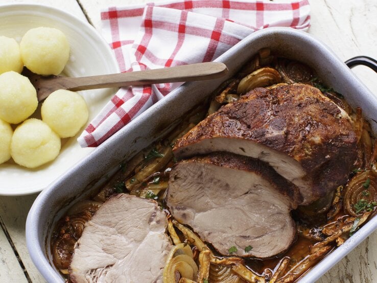 Bräter mit Schweinebraten in Soße, daneben ein Teller voll Knödel. 