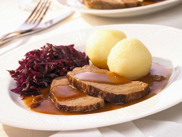 Rheinischer Sauerbraten mit Rotkohl und Kartoffelklößen auf weißem Teller. 