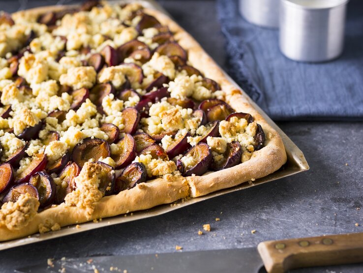 Ein silbernes Blech mit dem zuckerfreien Zwetschgenkuchen auf einem dunkelblauem Untergrund von schräg oben fotografiert.