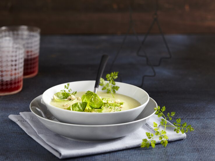 Die Rosenkohl-Cremesuppe in weißem Geschirr auf einem dunkelblauen Tisch und dunkelblauer Wand. Alles von der Seite fotografiert.