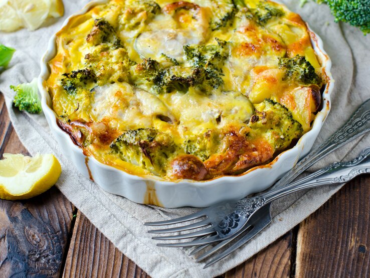 Eine weiße Auflaufform mit dem Gratin auf einem rustikalen Holztisch mit weißer Tischdecke.