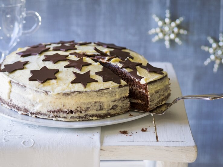 Weißer Kuchen mit schokoladigen Sternen vor weihnachtlich geschmücktem, blauen Hintergrund. 