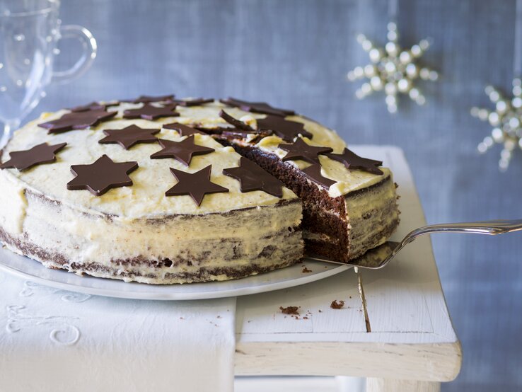 Weißer Kuchen mit schokoladigen Sternen vor weihnachtlich geschmücktem, blauen Hintergrund. 