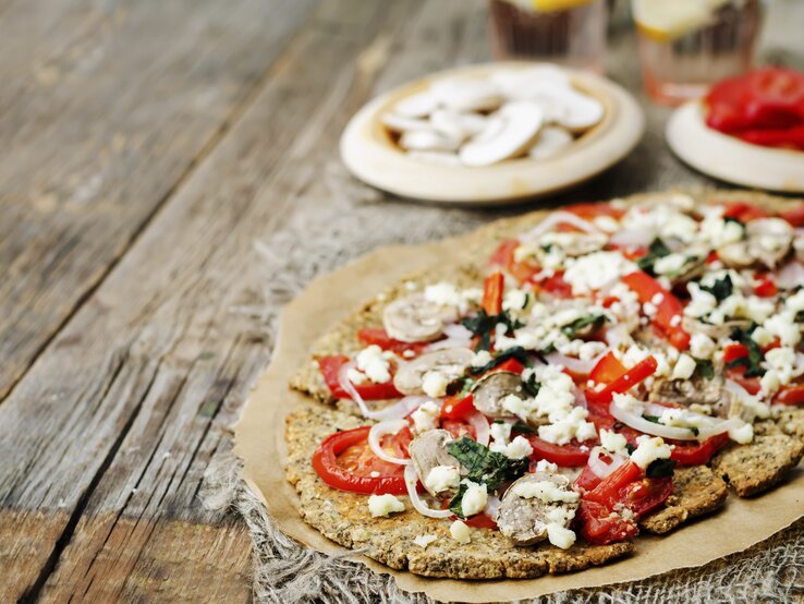 Die Chiasamen-Pizza auf einem rustikalem Holztisch von schräg oben fotografiert. Im Hintergrund noch unscharf Zutaten zum Belegen zu erkennen.