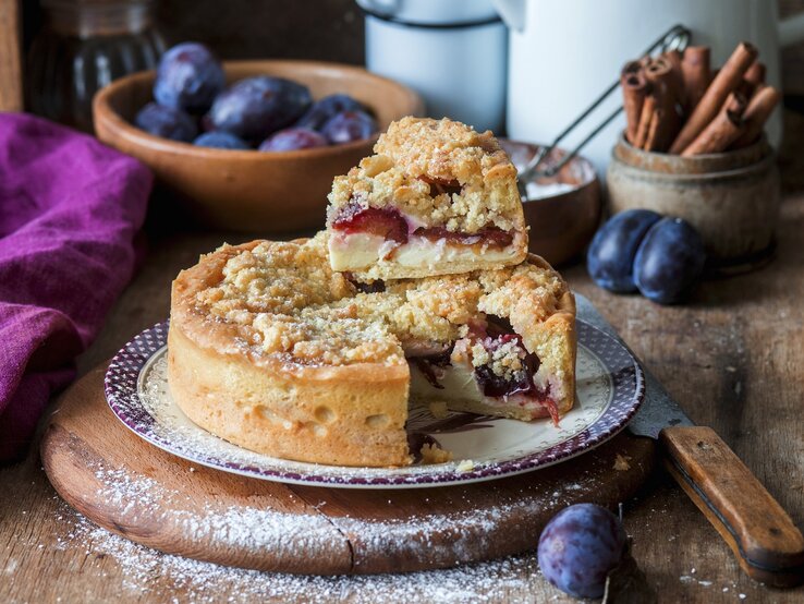 Der Streusel-Zwetschgen-Quarkkuchen auf einem rustikalen Holztisch mit Zwetschgen im Hintergrund fotografiert.