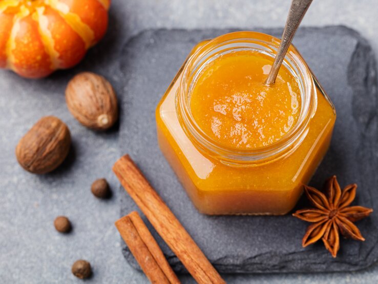 Ein Glas mit Kürbis-Orangen-Marmelade steht auf einer Steinplatte auf einem grauen Untergrund. Neben dem Glas liegt Anis und zwei Zimtstangen. Neben der Steinplatten liegen einige Nüsse und ein Zierkürbis. 