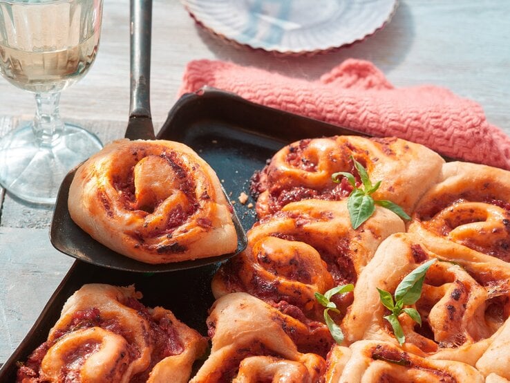 Pizza-Schnecken vom Blech auf einem schwarzen Blech auf hölzernem Hintergrund. Links ein Glas Sekt.