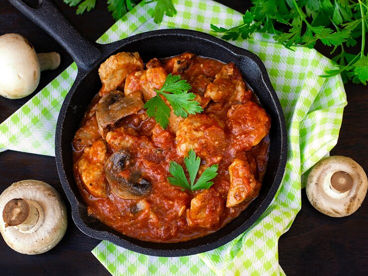 Eine gusseiserne Pfanne mit Tomatensauce, Pilzen und Fleischstücken steht auf einem karierten Tuch, umgeben von frischen Champignons.