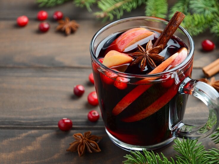 Eine durchsichtige Tasse gefüllt mit selbst gemachtem Glühwein auf Holztisch. Daneben weihnachtliche Gewürze und Deko-Elemente. 