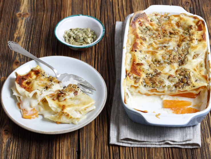 Die vegetarische Kürbis-Lasagne mit Kürbiskernen auf einem dunklen Holztisch von schräg oben fotografiert.