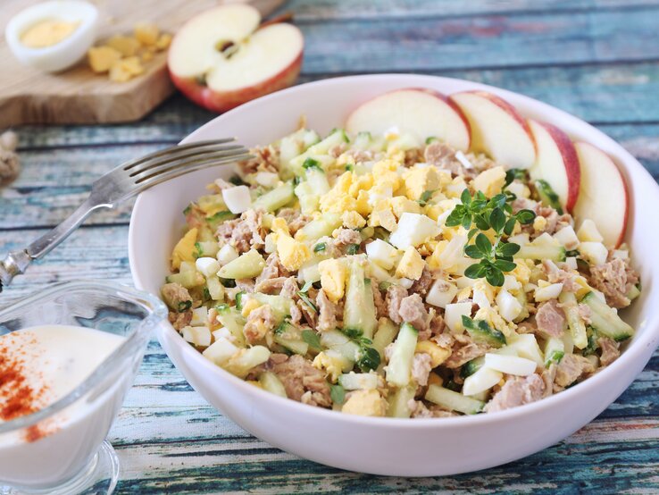 Cremiger Salat aus Ei, Thunfisch und Gurken, mit Apfelscheiben und Kräutern garniert, serviert auf einem Holztisch.