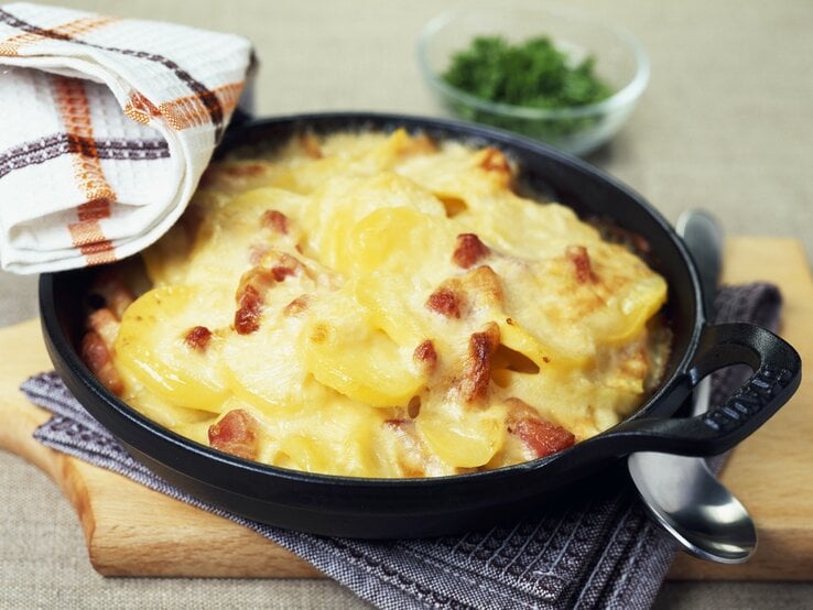 Kartoffelauflauf mit Schinken und Käse in gusseisernen Form auf Holzbrett.