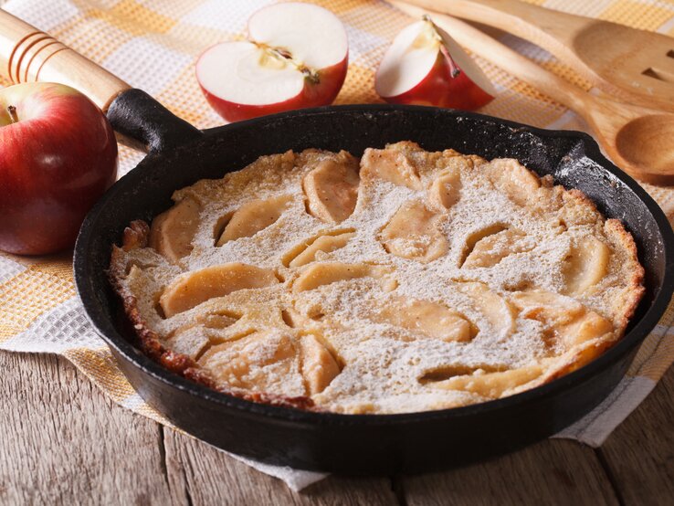 Apfelpfannkuchen in gusseiserner Pfanne auf gelb-weiß karriertem Tischtuch.