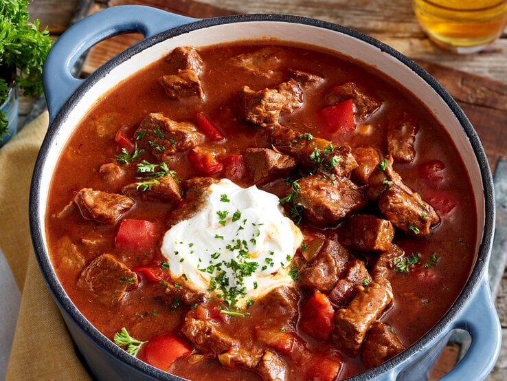 Brauhaus-Gulasch mit Kartoffel-Pastinaken-Stampf und Schmand in einem Topf mit blauen Griffen auf Holzbrett und Holztisch. Im Bild ein Glas mit Petersilie und ein Glas Bier angedeutet. Draufsicht.