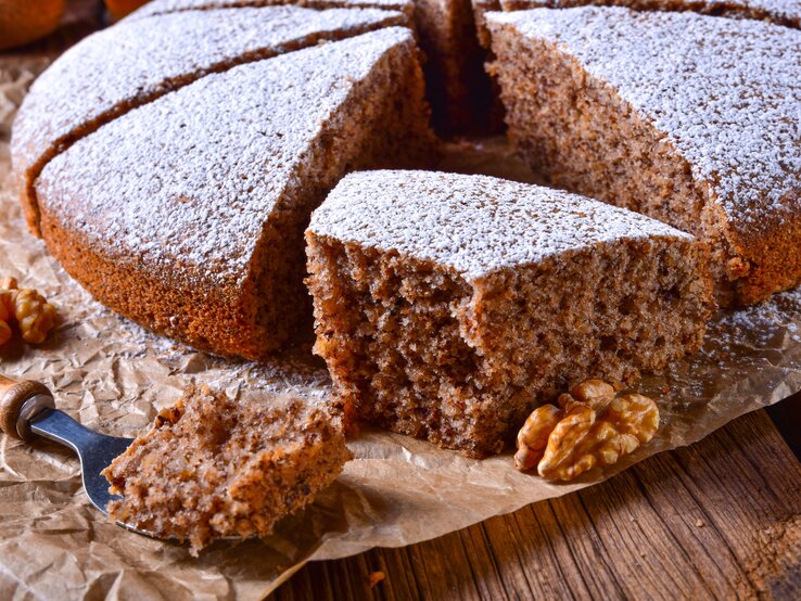 Ein angeschnittener Walnusskuchen mit Schokolade, bestäubt mit Puderzucker, auf einem Holztisch mit Backpapier. 