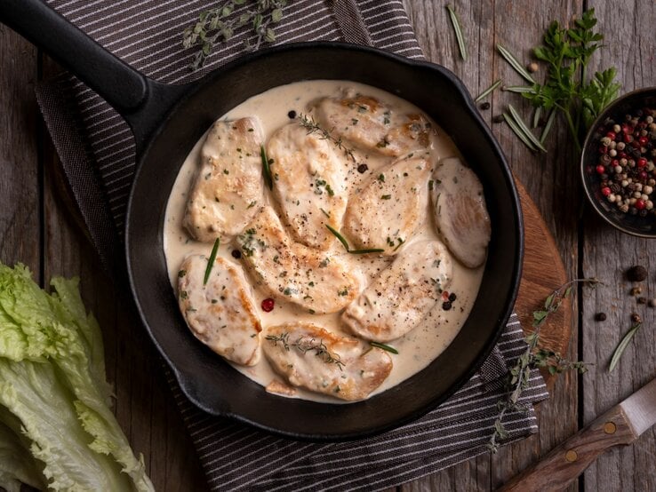 Hähnchen in mexikanischer Sahnesoße in Gusspfanne auf Tischtuch und Holztisch. Im Bild eine Schale mit buntem Pfeffer, Kräutern und Salat. Draufsicht.