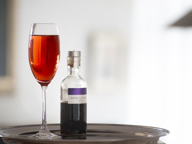 Ein Glas mit dem Kir Royal vor hellem Hintergrund und daneben eine Flasche mit dem Cassis-Sirup.