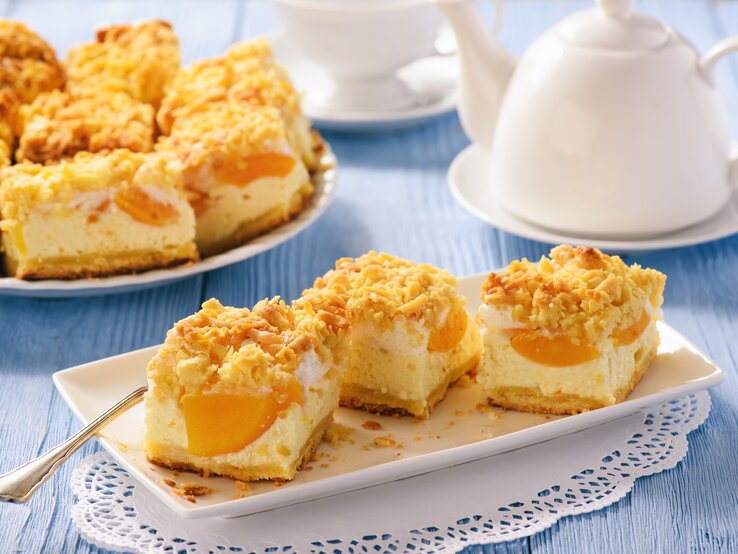 Geschnittener Pfirsich-Käsekuchen mit Streusel  auf weißem Geschirr auf blauer, hölzerner Oberfläche.