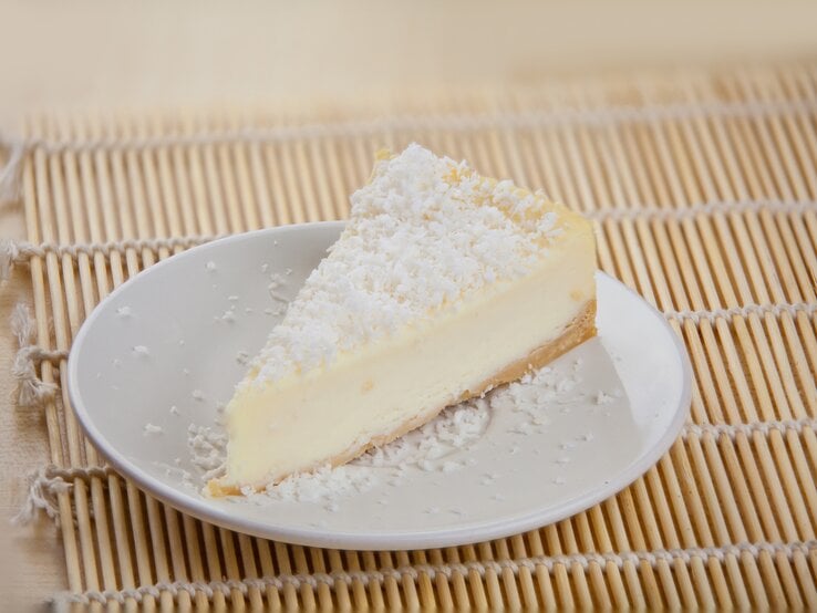 Ein Stück cremiger Joghurtkuchen mit Kokosraspeln bestreut auf einem weißen Teller, der auf einer Bambusmatte steht.