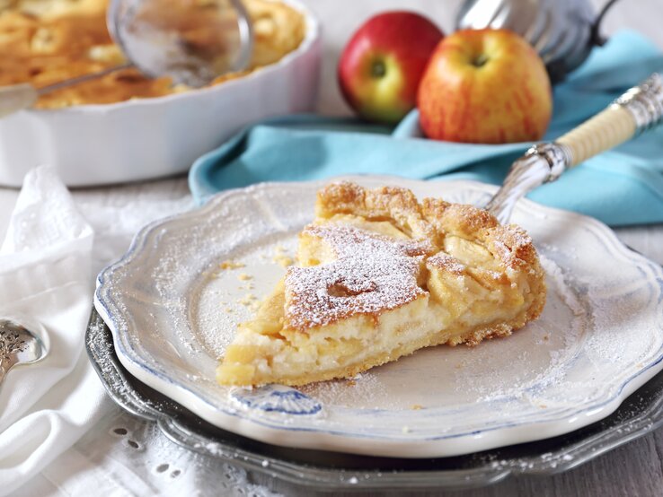 Ein Stück Apfelkuchen mit Puderzucker bestäubt liegt auf einem Teller, im Hintergrund ein ganzer Kuchen und zwei Äpfel.