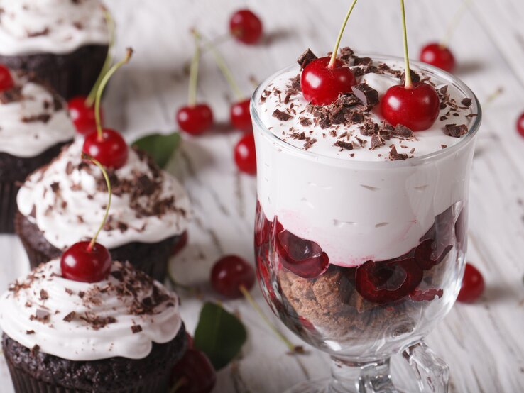 Schoko-Sahne-Kirsch-Dessert im Glas umrundet von Schokomuffins auf hölzernem Untergrund.