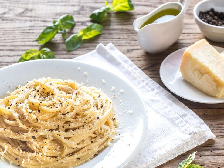 Ein Teller Pasta Cacio e Pepe auf einem rustikalen Holztisch mit Zutaten wie Pfeffer und Parmesan. Alles von schräg oben fotografiert.