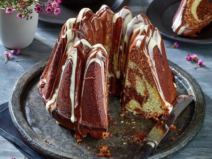 Ein saftiger Marmorkuchen mit Schokoladenglasur und heller Verzierung steht auf einem silbernen Teller, ein Stück wurde herausgeschnitten.