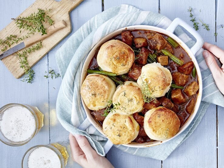 Bier-Gulasch mit Brötchen in einem weißen Topf auf hellem Hintergrund.