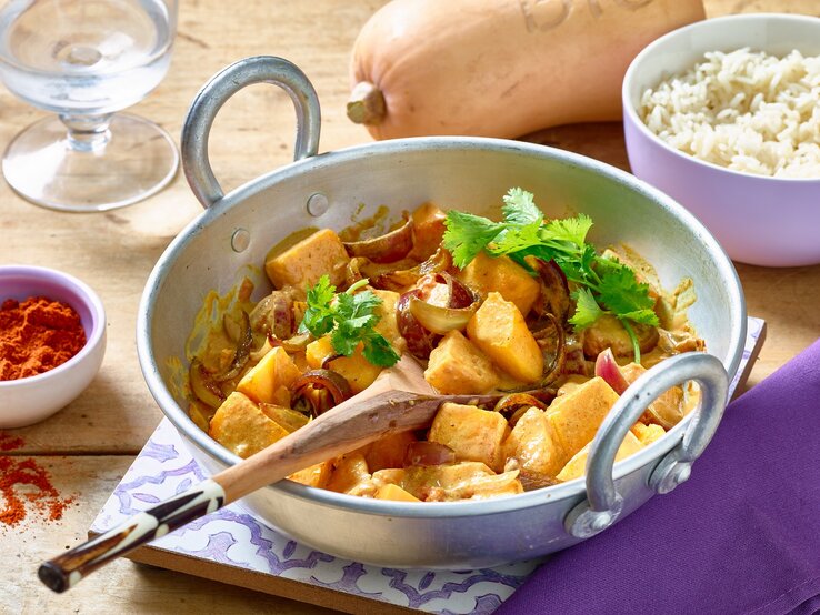 Kürbis-Curry in einer weißen Pfanne auf hölzernem Hintergrund. Rechts eine Schüssel mit Reis, links Gewürze und ein Wasserglas. Oben auf dem Foto ein Kürbis.