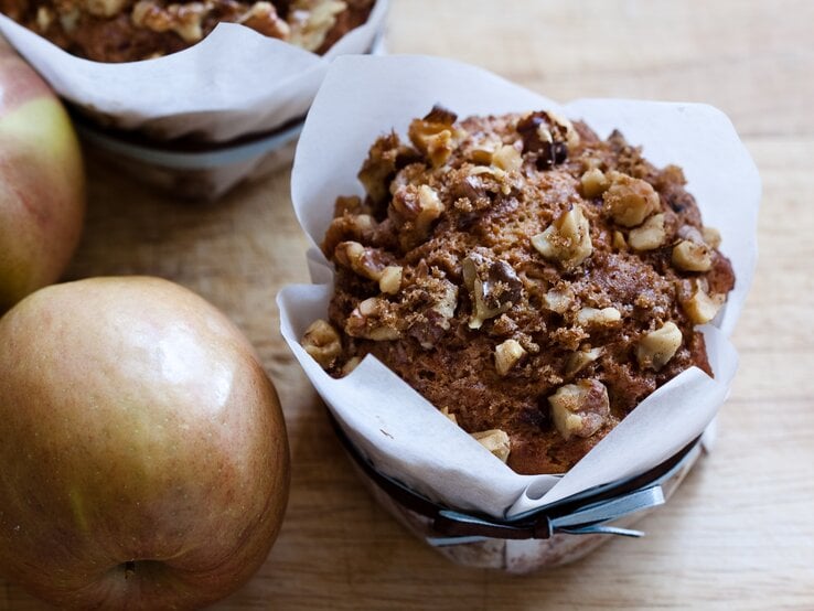 Ein saftiger Apfel-Walnuss-Muffin in weißem Papier liegt auf einem Holztisch, daneben zwei frische, rote Äpfel.