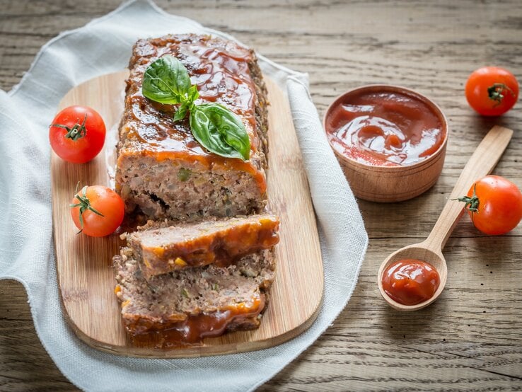 Ein Lieblingshackbraten mit Tomaten-Rotwein-Überzug liegt auf einem Holzbrett auf einem Geschirrtuch. Um ihn  herum liegen ein Löffel, Tomaten und eine Schale mit Tomaten-Rotwein-Mischung.