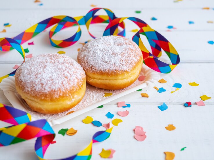 zwei Berliner mit Puderzucker bestäubt in weißem Szenario. Daneben Konfetti und Luftschlangen.