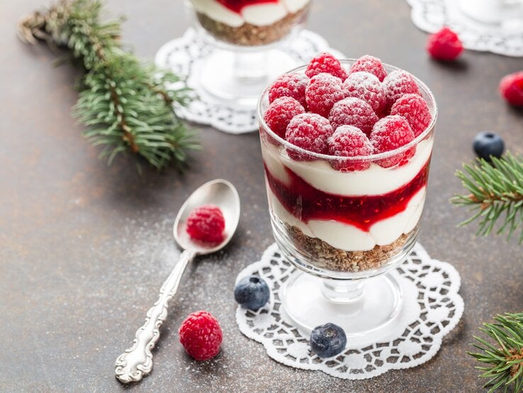 Dessert im Glas mit Himbeeren und Creme mit Kuchenboden auf dunklem Untergrund. Umgeben von Tannenzweigen.