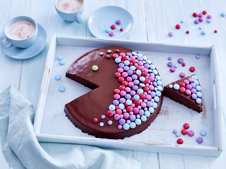 Ein Schokoladen-Kuchen in Fischform der mit bunten Streuseln beleget ist auf einem hellblauen Untergrund und weißem Tablett von oben fotografiert.