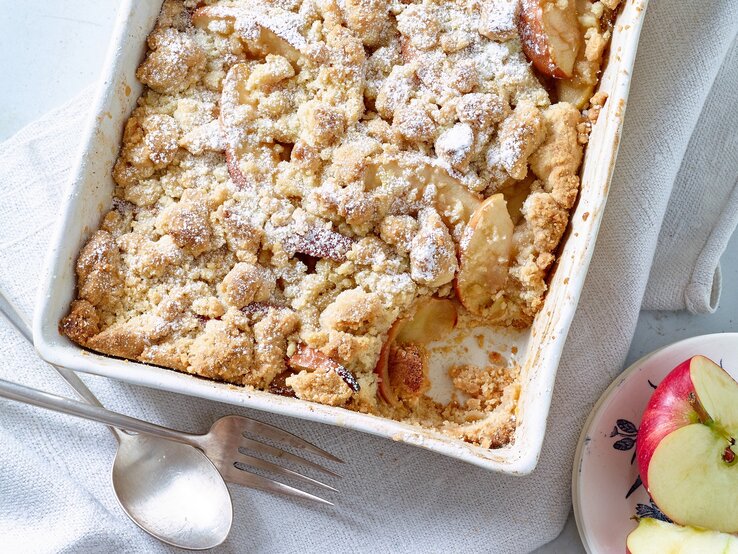 Apple Crumble auf weißem Tuch, daneben Äpfel und Besteck.