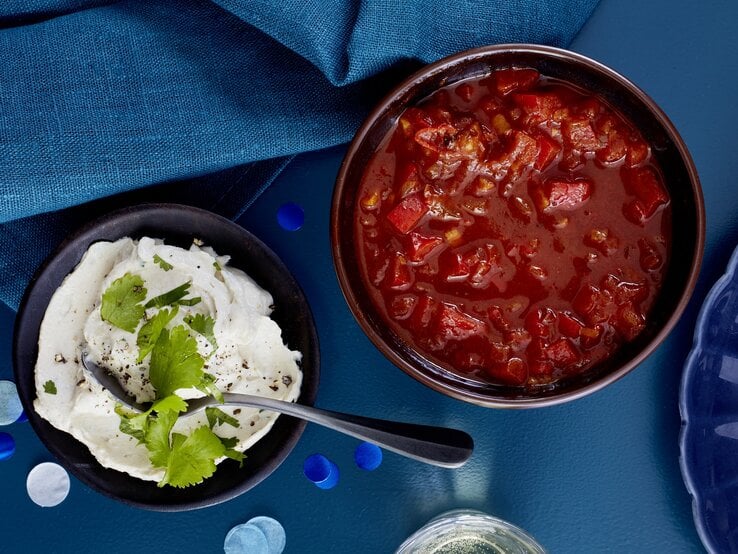 Paprika-Tomaten-Salsa in einer Schüssel, links daneben ein weiterer Dip. Der Hintergrund ist dunkelblau und darauf verteilt ein dunkelblaues Geschirrtuch.