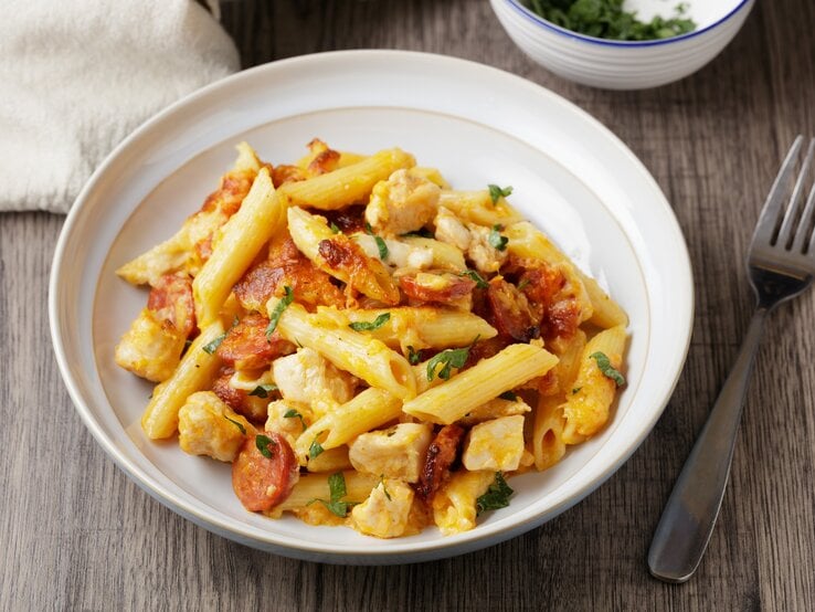 Pasta mit Bärlauch, Chorizo und Hähnchenbrust in weißem Pastateller auf Holz. Daneben eine Schale mit Bärlaich, ein Tuch und eine Gabel. Draufsicht.