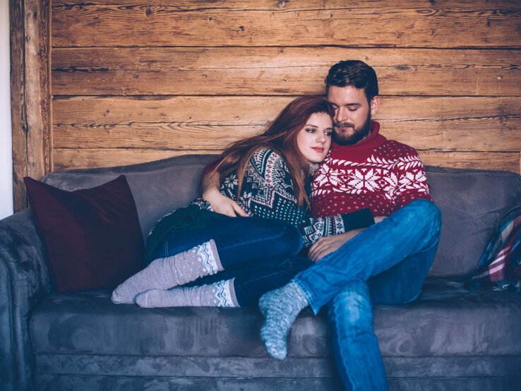 Paar, das gemütlich auf einem Sofa sitzt und sich eng aneinander kuschelt. Beide tragen warme Winterpullover mit skandinavischem Muster.