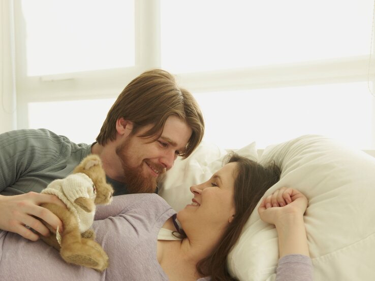 Ein lächelnder Mann mit braunem Bart und eine dunkelhaarige Frau in lila Oberteil liegen glücklich im Bett, mit einem Stofftier in seiner Hand.