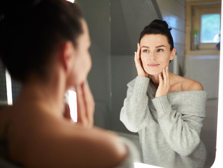 Junge Frau, die vor einem Spiegel in einem modernen Badezimmer steht. Sie trägt einen grauen, schulterfreien Pullover und hat ihr Haar zu einem eleganten Dutt hochgesteckt. Mit beiden Händen berührt sie sanft ihr Gesicht. | © iStock/shironosov