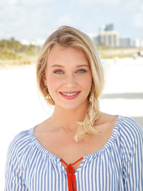 Junge Frau mit blonden Haaren und strahlend blauen Augen am Strand. Sie lächelt freundlich in die Kamera und trägt ein gestreiftes, blau-weißes Oberteil. Ihre Haare sind zu einem lässigen Zopf gebunden | © Bridget Cramer