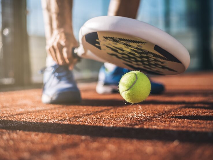 Eine Person in blauen Sportschuhen bereitet sich darauf vor, mit einem Padelschläger einen gelben Tennisball auf einem sandigen Platz zu schlagen.