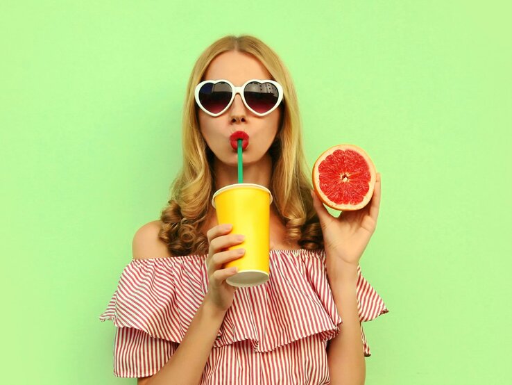 Eine blonde Frau vor grünem Hintergrund. Sie trägt eine Sonnenbrille, hält eine aufgeschnitte Grapefruit hoch und trinkt mit einem Strohhalm aus einem Becher.