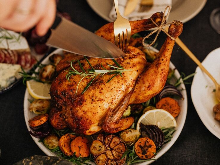 deutsche-kueche-brathaehnchen.jpg Ein goldbraun gebratenes ganzes Hähnchen wird auf einem Teller mit Ofengemüse, Zitronenscheiben, Knoblauch und frischen Kräutern serviert und gerade angeschnitten.